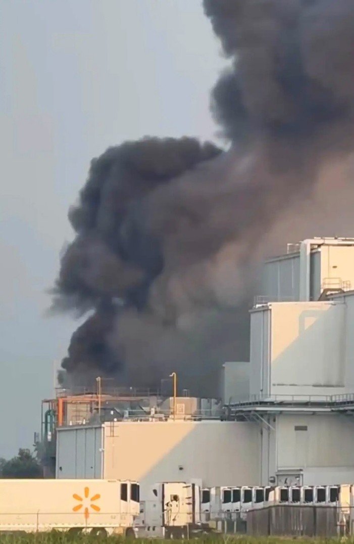 Plumes of heavy black smoke pouring from an industrial facility fire above parked semi-trailers.