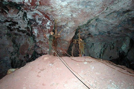 Two miners operating drilling equipment set up on vertical support poles inside a dark underground hard rock stope, standing atop a mound of reddish broken ore.