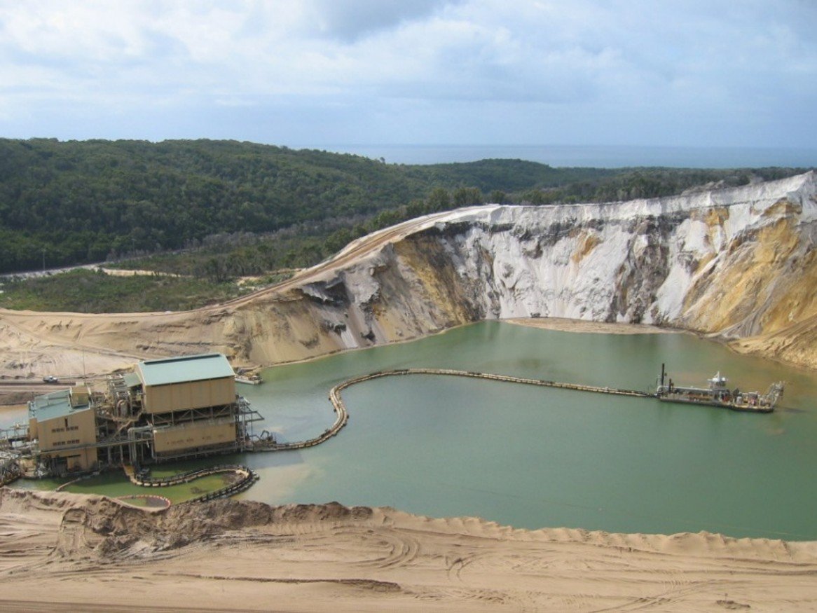 High-angle view of an Australian titanium sand mining operation featuring a floating dredge on a green pit pond connected by a long floating pipeline to a large processing plant.