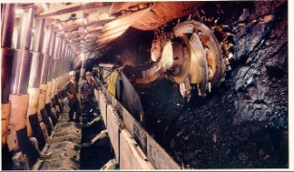 Two miners standing beside a massive longwall shearer machine with a large rotating cutter drum cutting into a dark coal face, supported by heavy hydraulic roof supports.