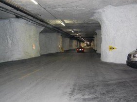 A wide, brightly lit underground room-and-pillar mine tunnel with white rock walls, paved floors, and vehicles driving along the passage.