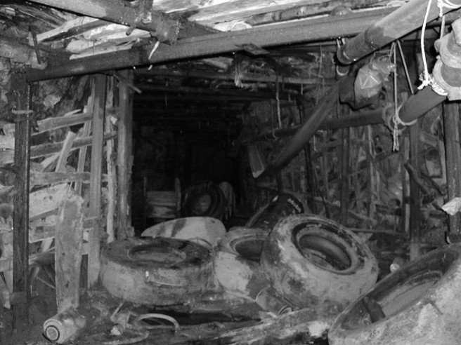 Black and white photo of a dark, cluttered underground mine shaft filled with discarded rubber tires, wooden timber sets, pipes, and old equipment.
