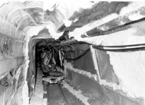 Black and white photo of a narrow underground mine tunnel lined with flexible plastic ventilation curtains along the walls and ceiling, with rail tracks running along the floor.