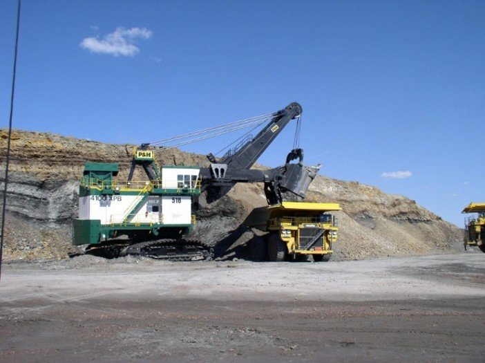Large P&H electric rope shovel filling the bed of a giant yellow haul truck in an open-pit mine under a clear blue sky.
