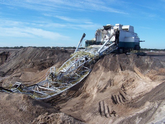 Collapsed white lattice boom of a massive dragline excavator resting broken against dirt embankments at a coal mine.