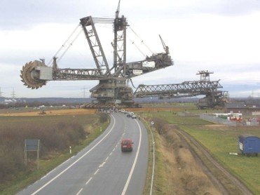 A massive bucket-wheel excavator crossing a two-lane paved road in Germany, dwarfing the vehicles on the highway and nearby buildings.