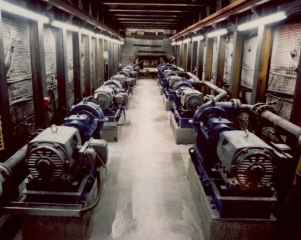 Two rows of heavy-duty blue electric water pumps lined up inside a bright, narrow underground mine corridor.