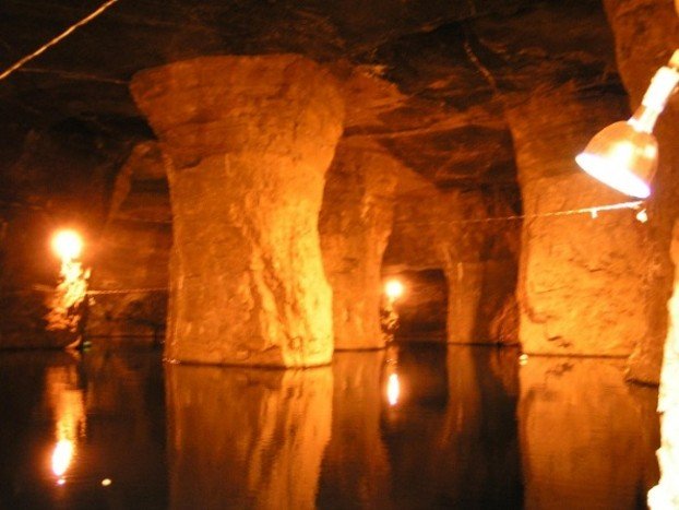 Underground room-and-pillar mine chamber flooded with still, dark water reflecting orange lights and large supporting rock pillars.