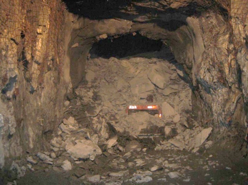 Underground mine tunnel blocked by a heavy pile of collapsed roof rock, partially burying a piece of mobile equipment with its orange tail lights glowing.